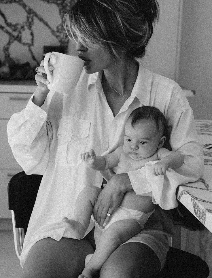 A mother in an oversized shirt drinking coffee, baby on her lap
